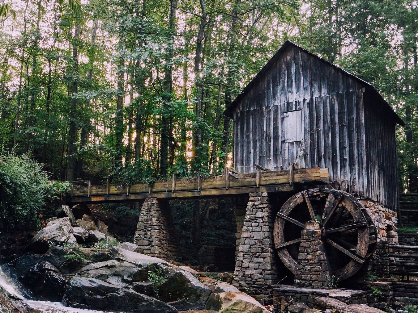 A water mill in the middle of a forest with a waterfall in the background.