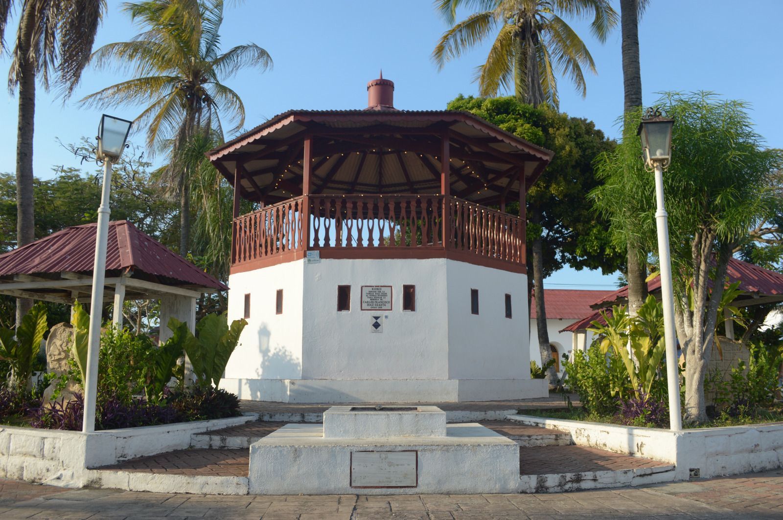 White and brown gazebo in a plaza with palm trees under a clear blue sky.