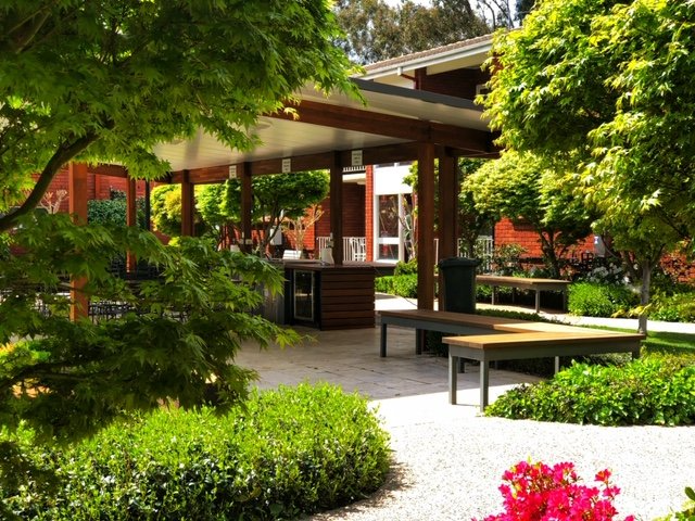 Outdoor patio with wooden beams, benches, and landscaping; gravel ground, trees, and red building.