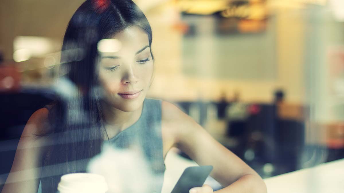 A woman is sitting in front of a window looking at her cell phone.