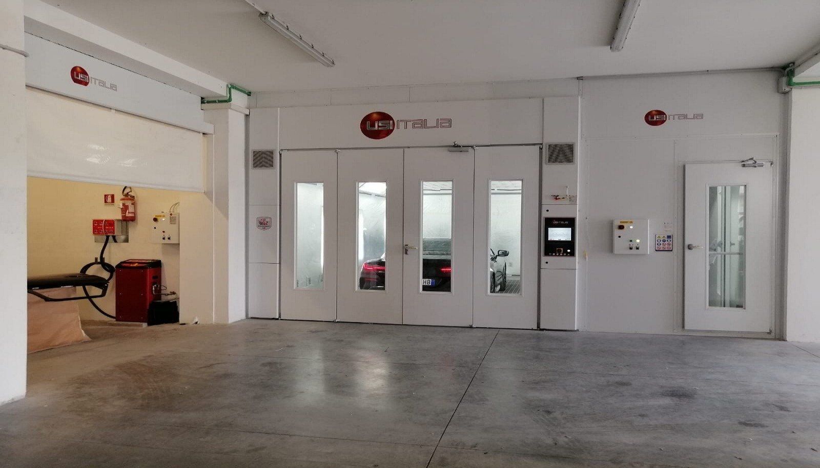 Interno di un'officina con cabina di verniciatura bianca e porte chiuse. Pavimento in cemento grigio e luci fluorescenti.