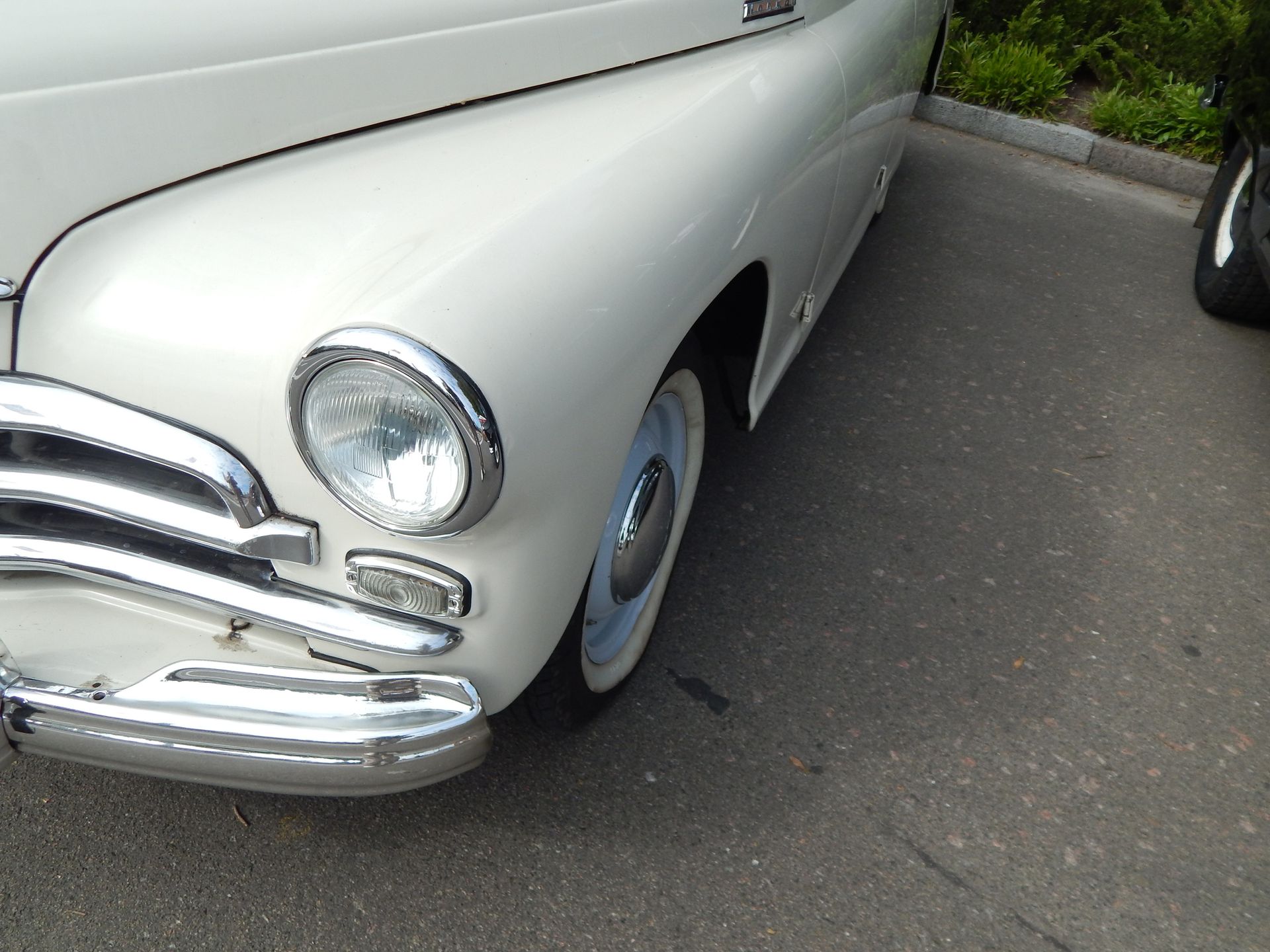 Cream-colored vintage car front with chrome details and white-walled tires parked on asphalt.