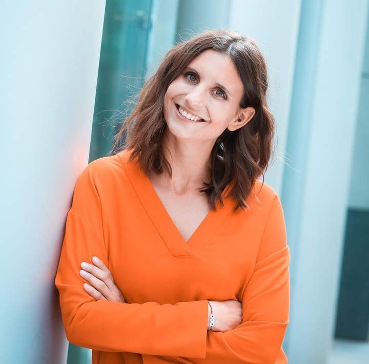 Eine Frau in einem orangefarbenen Hemd lehnt mit verschränkten Armen an einer Wand und lächelt.