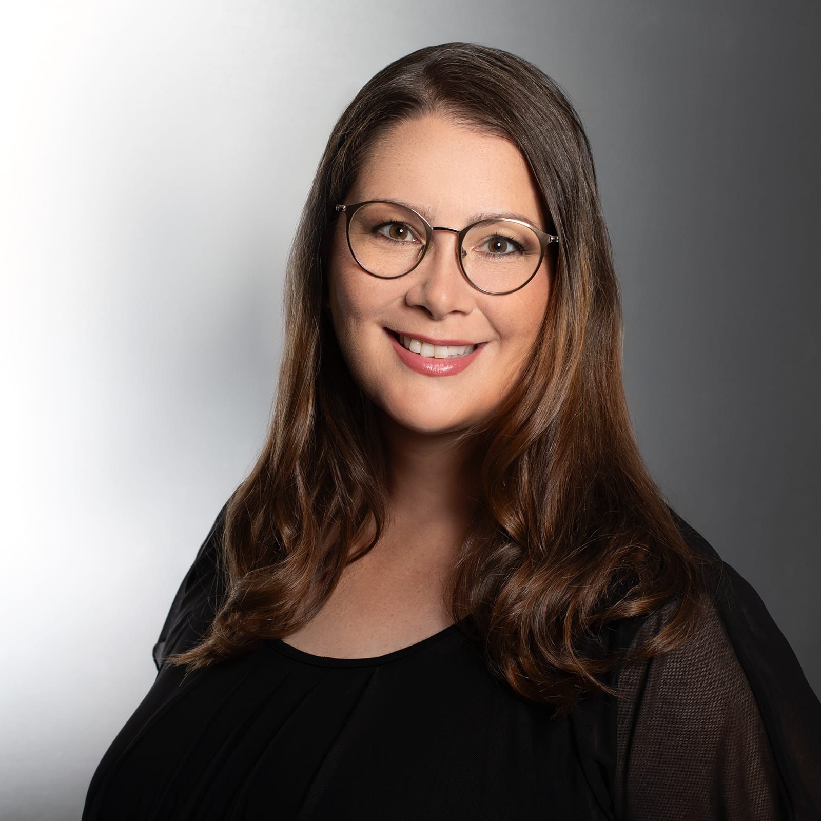 Eine Frau mit Brille und Halskette lächelt vor einer Backsteinmauer.