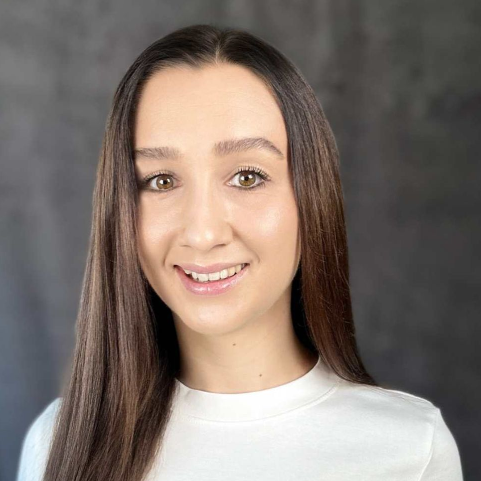 Frau mit langen braunen Haaren lächelt in die Kamera, sie trägt ein weißes Hemd, vor grauem Hintergrund.