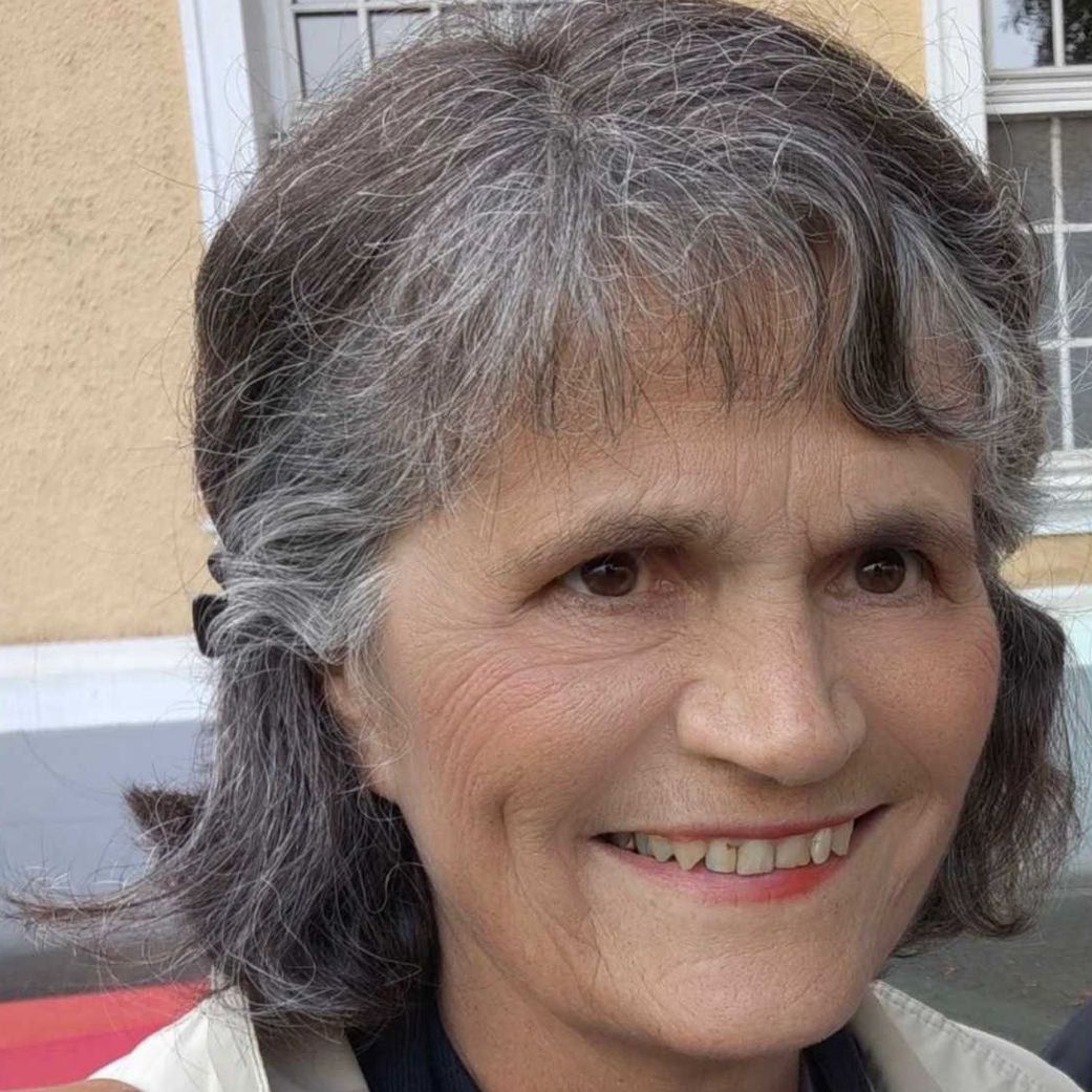 Eine Frau mit grauem Haar lächelt vor einem gelben Gebäude.