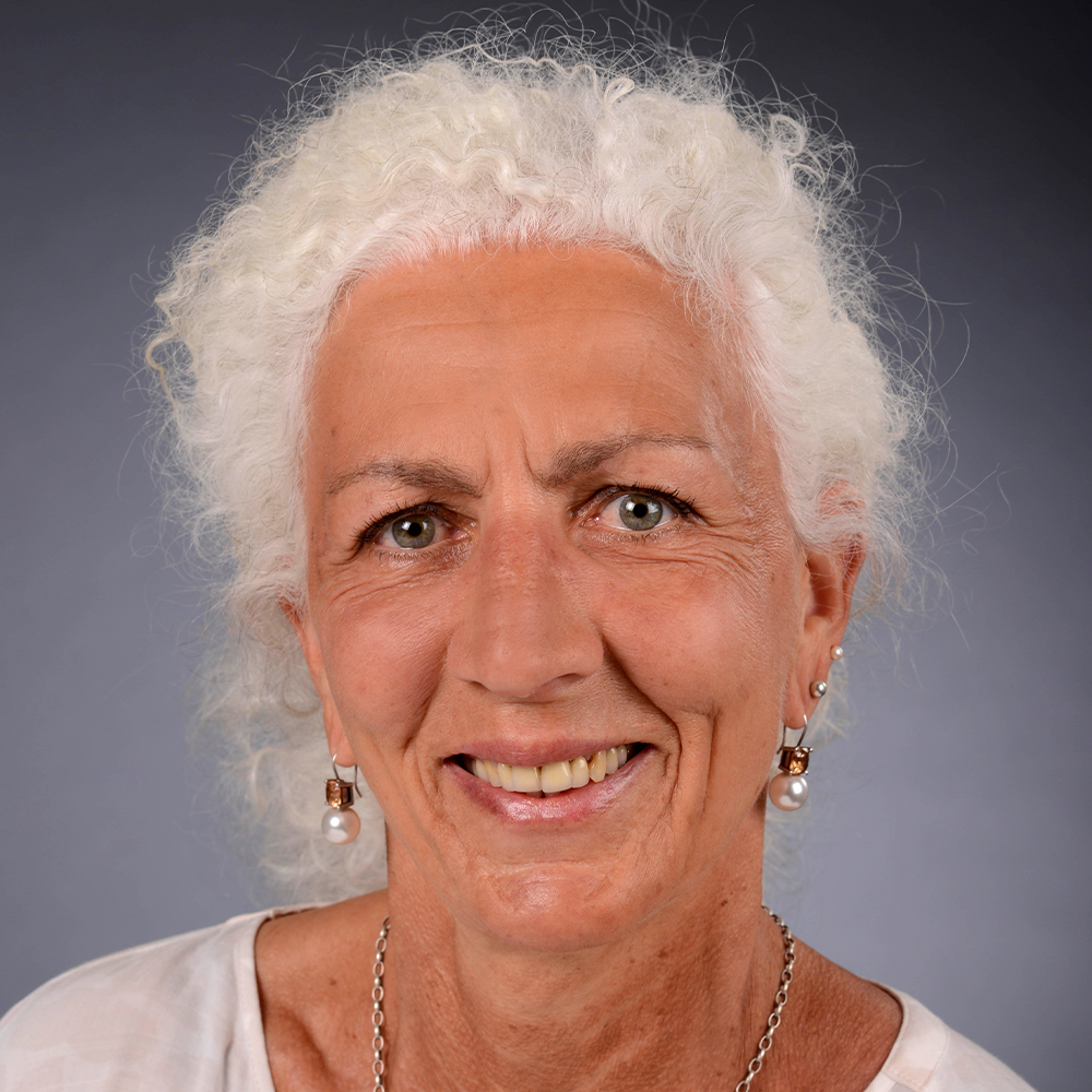 Eine Frau mit weißem Haar und Perlenohrringen lächelt in die Kamera.