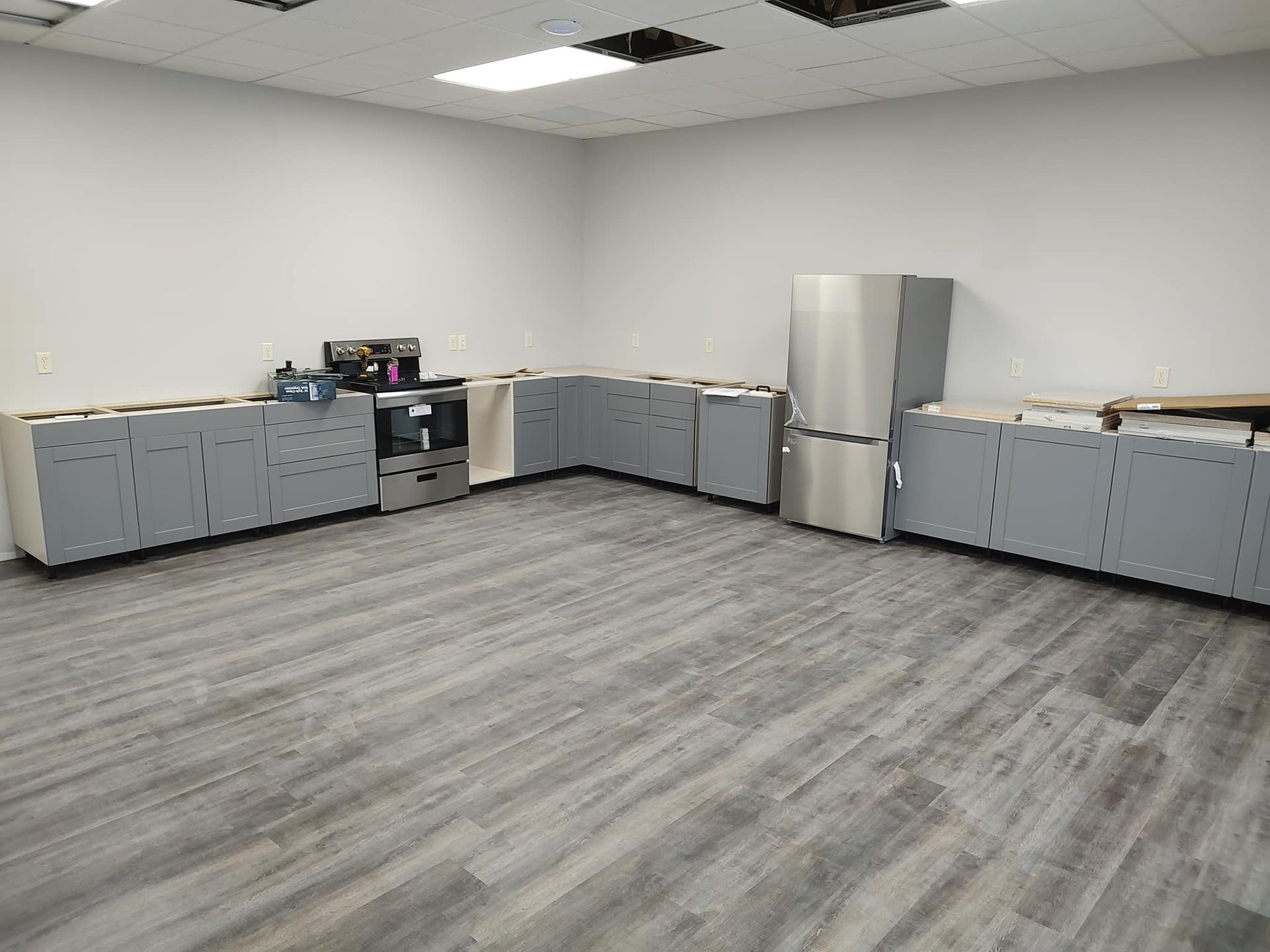 Gray kitchen cabinets and appliances arranged in an L-shape on gray wood-look flooring, in an empty room.