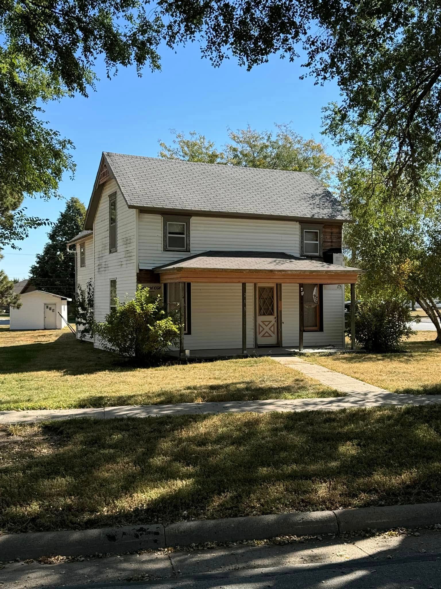 Two-story white house with porch and aging roof, trees on either side.