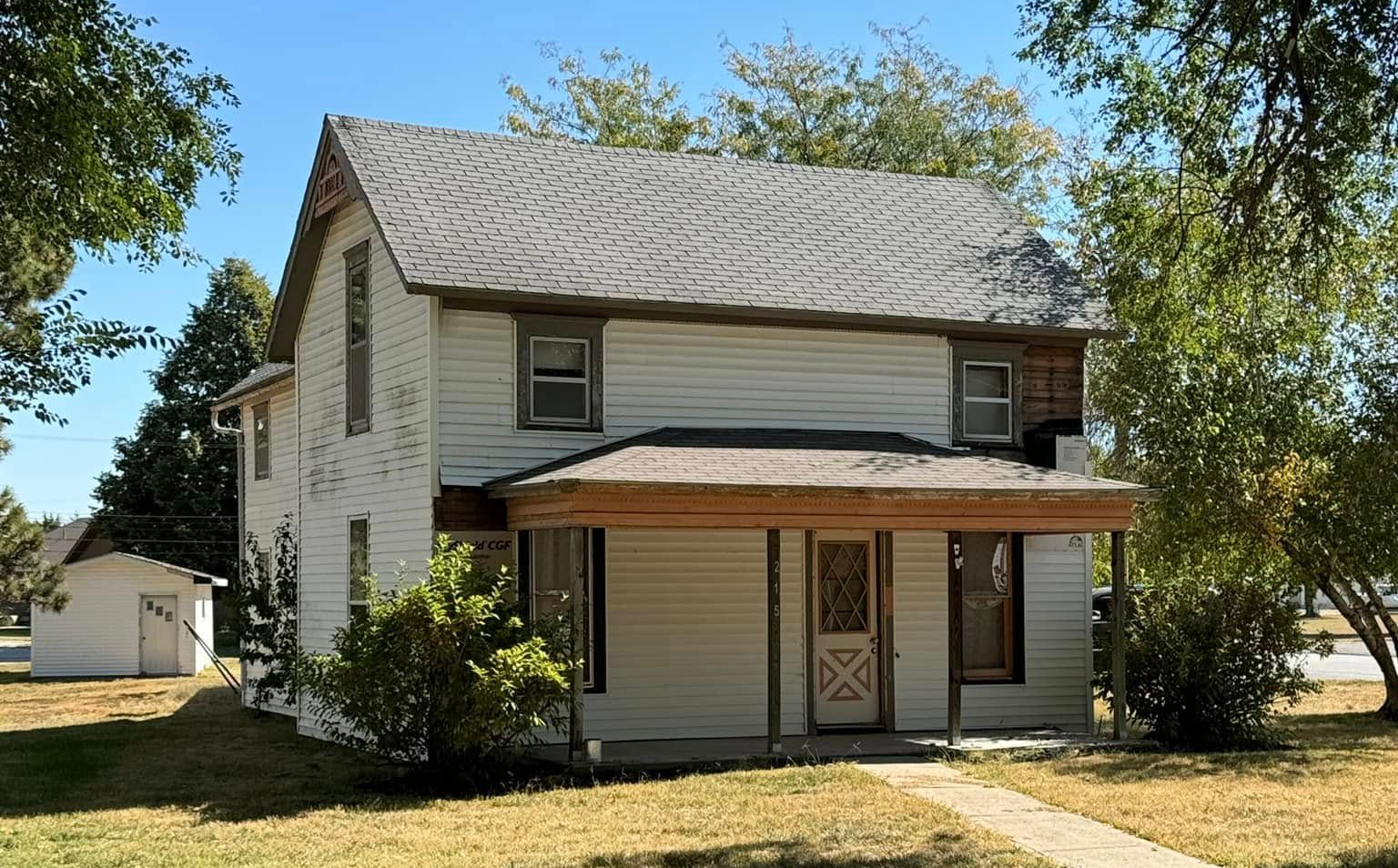 Two-story white house with brown trim and porch, on a grassy lot with trees.