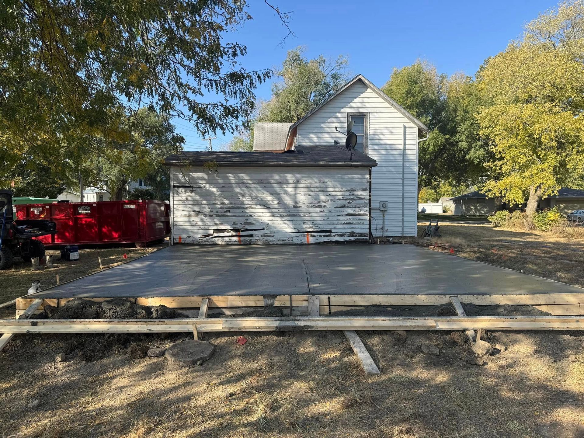 Newly poured concrete slab in front of a white building with peeling paint. Brown grass and trees in background.