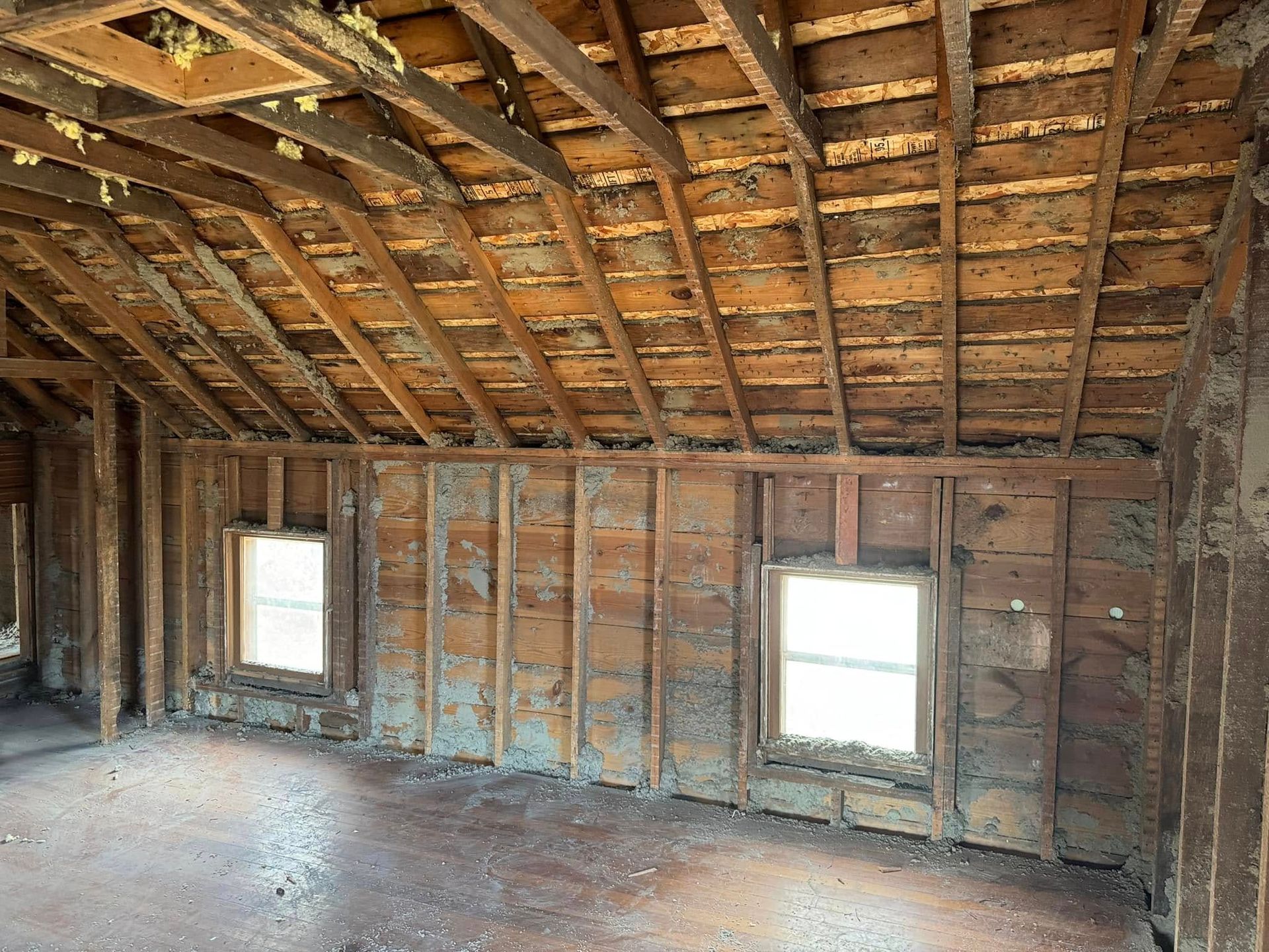 Interior view of a room under renovation; exposed wooden studs, ceiling beams, two windows, dirty floors.