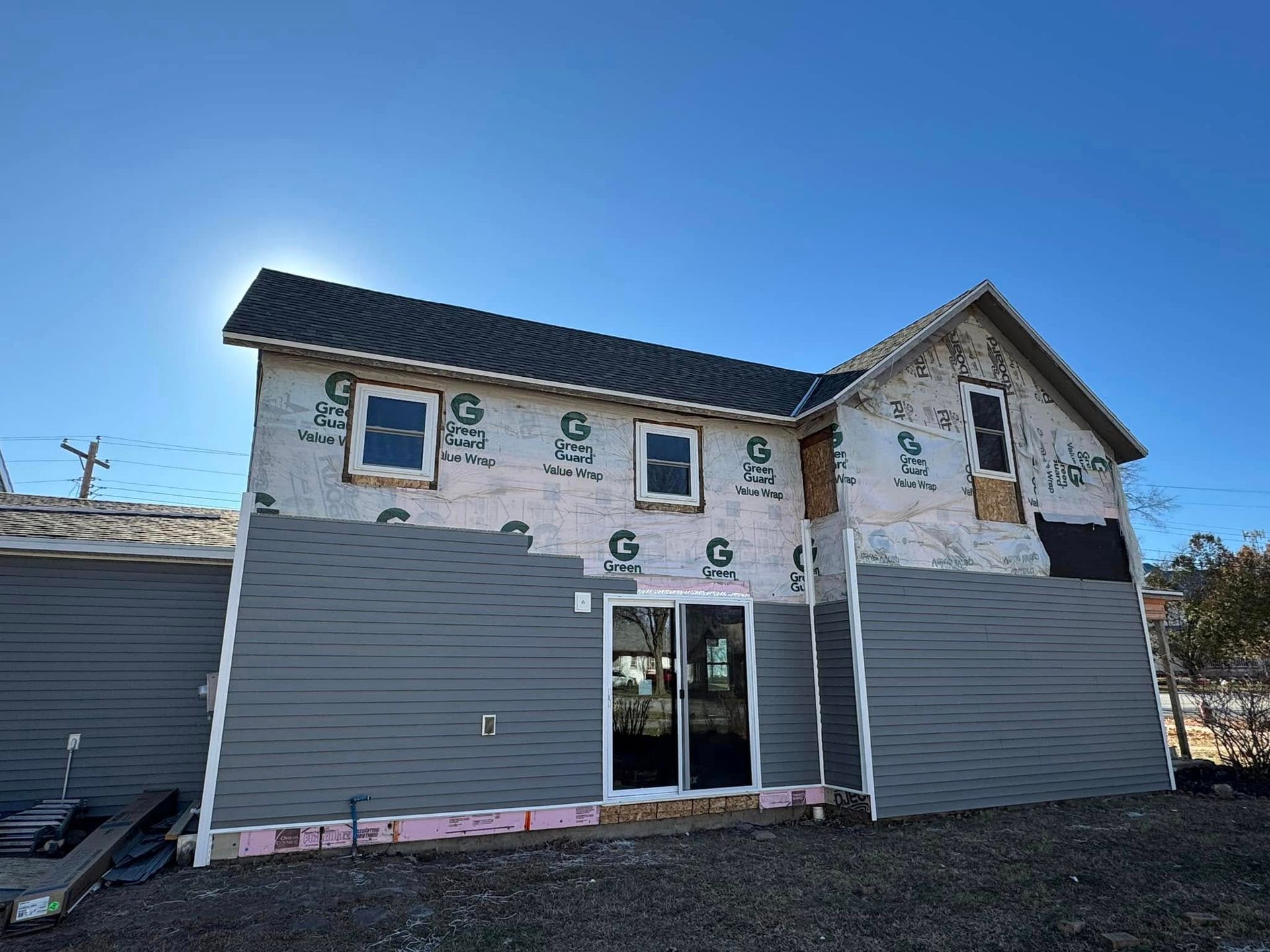 Building under construction; gray siding, windows, and tarpaper against a blue sky.