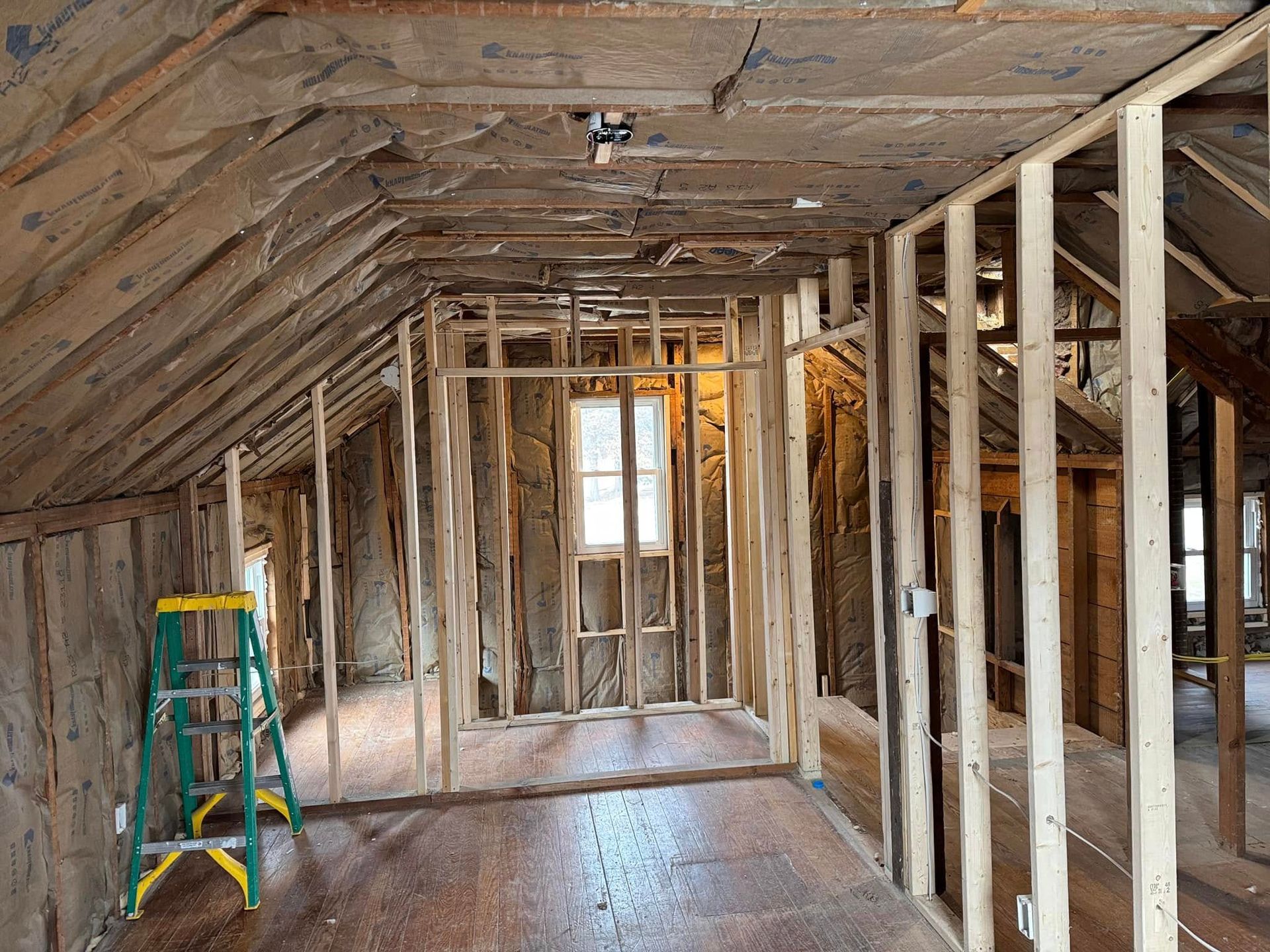 Interior of a room under construction with exposed wood framing, insulation, and a window. A ladder is present.
