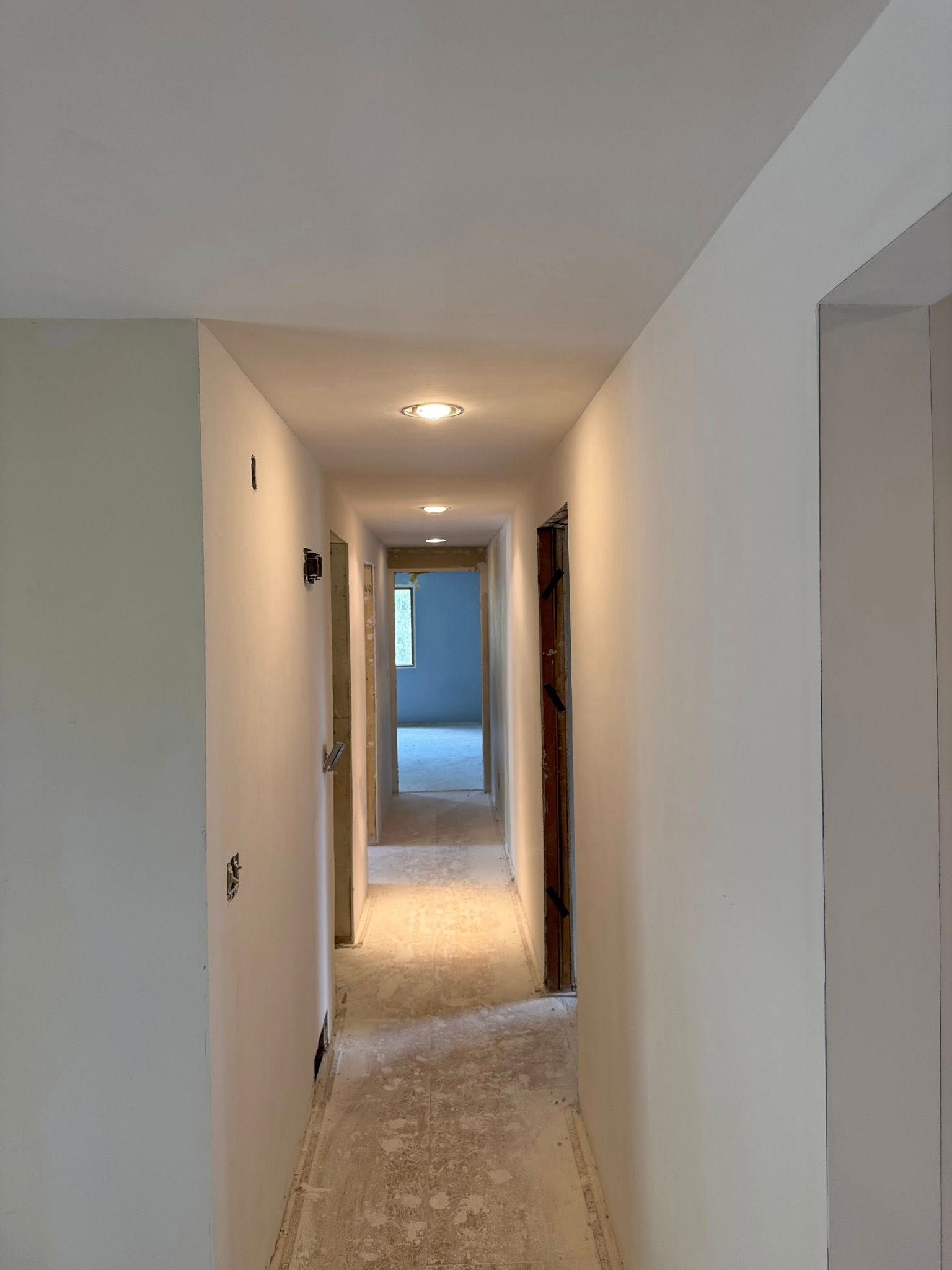 Narrow hallway with white walls and ceiling, doors on both sides, and recessed lighting.