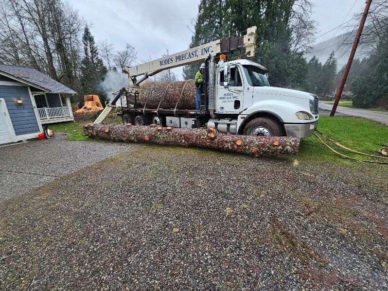 Truck with a crane loading logs; residential setting. Worker in cab. Pile of logs on the ground.