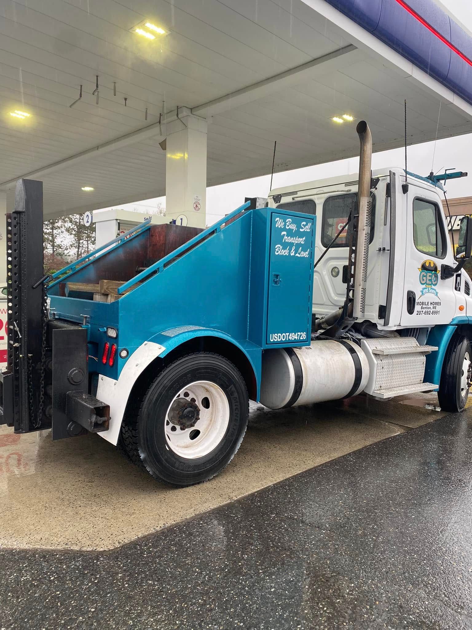 Truck with blue and white paint at a gas station under a canopy in the rain.