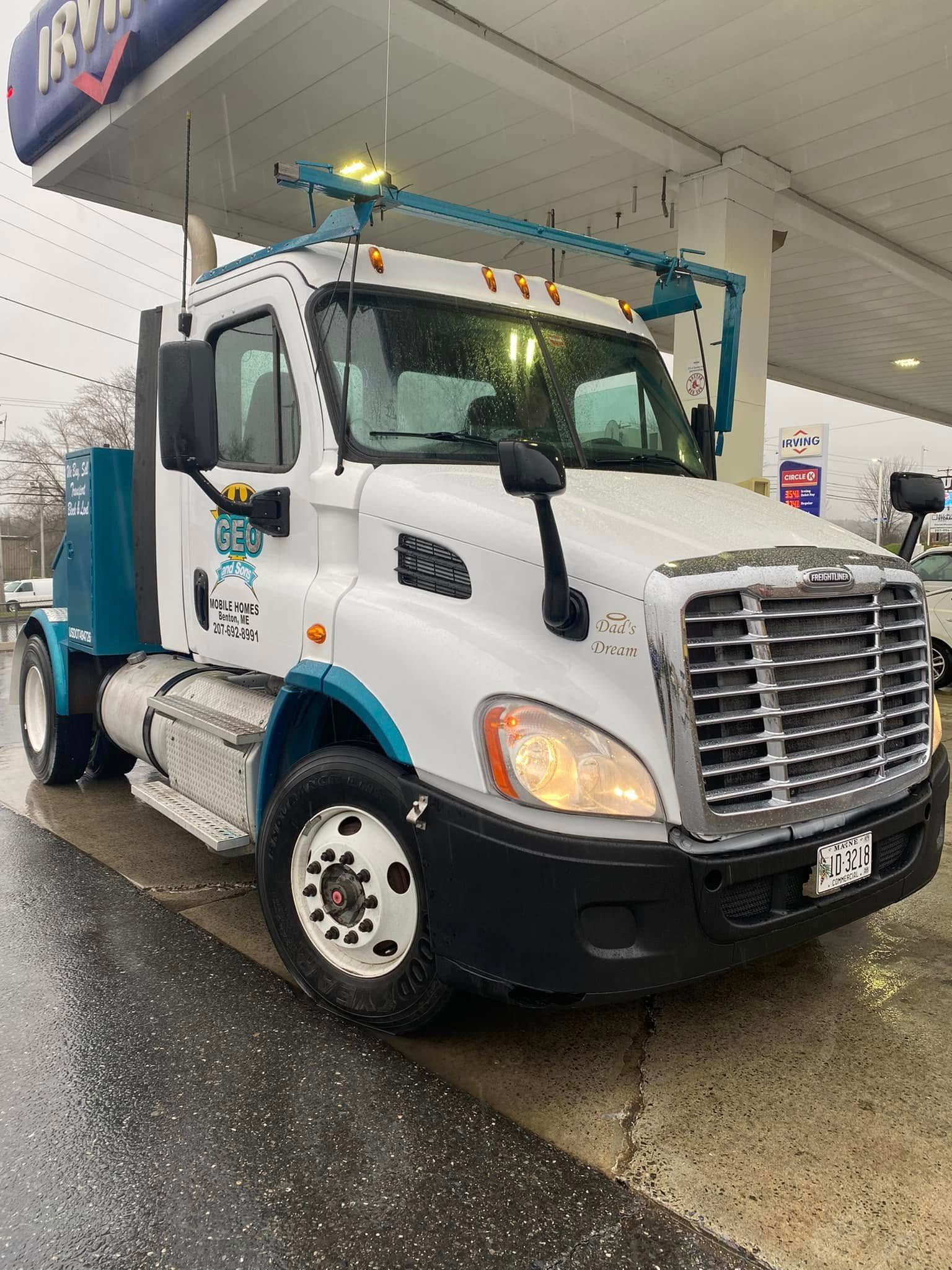 White truck with blue accents at a gas station under overcast skies; rain on the ground.