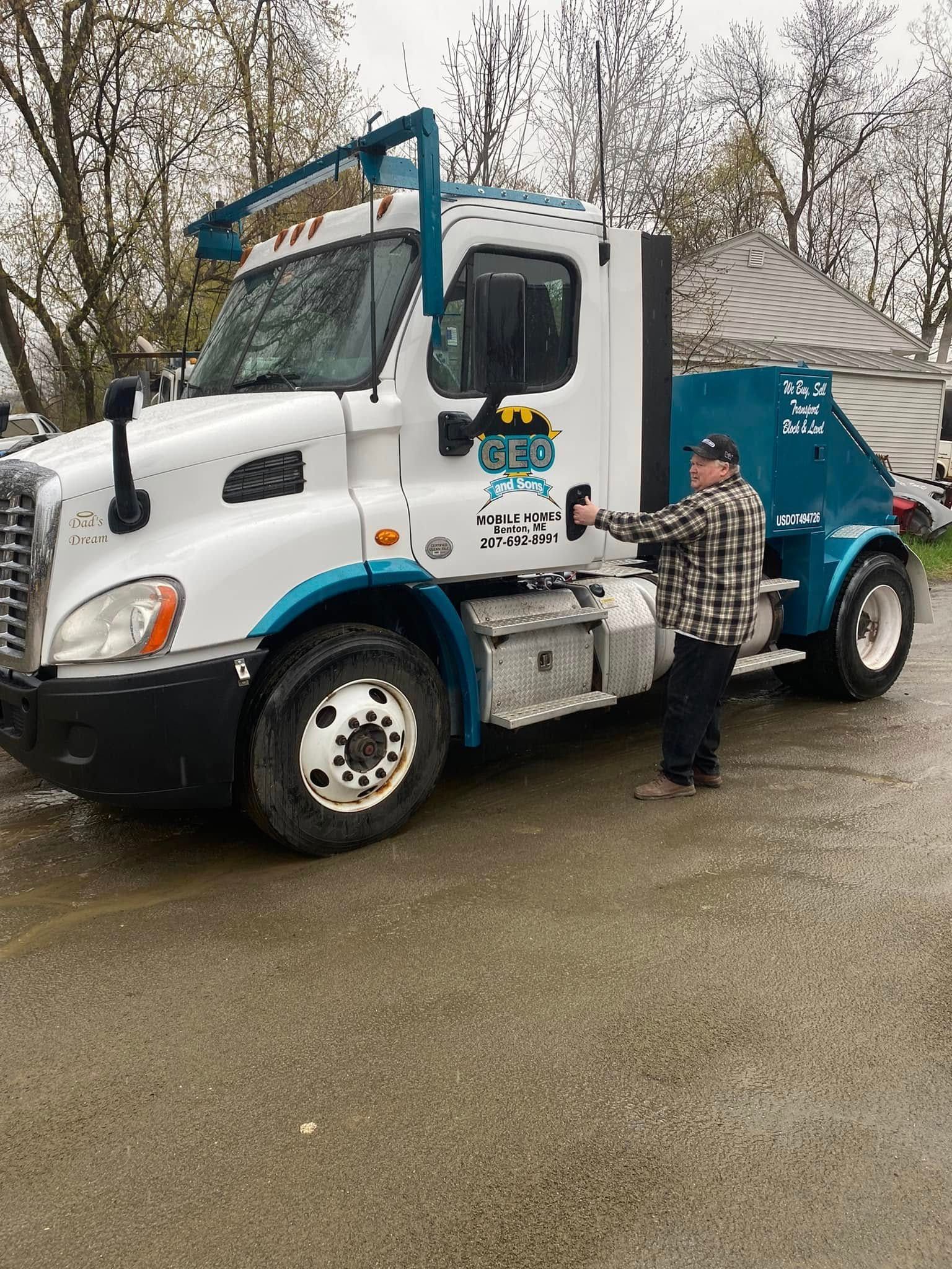 Man standing by a white and teal semi-truck with logo on door; cloudy day.