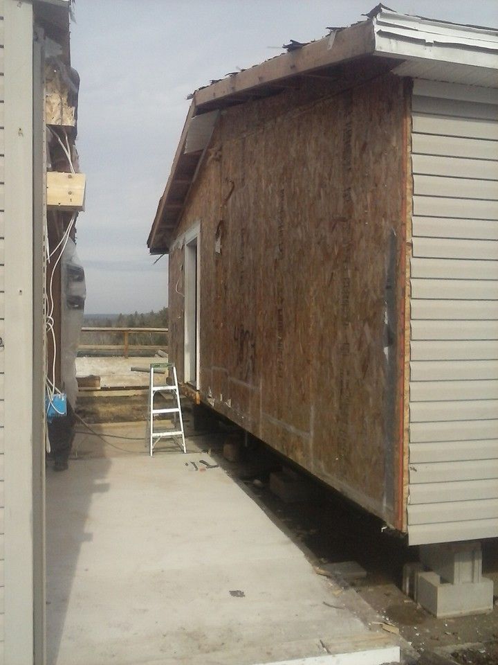 Side view of a building under construction, showing plywood walls and gray siding, with a small ladder and concrete walkway.