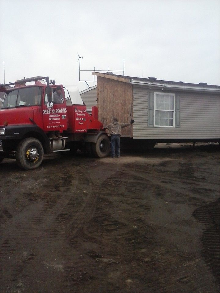 Red truck towing a mobile home on muddy ground; person standing near the structure.