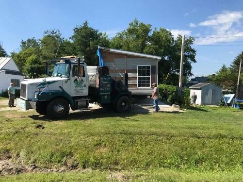 Truck transporting a small shed on a grassy area, blue sky. Two people nearby.