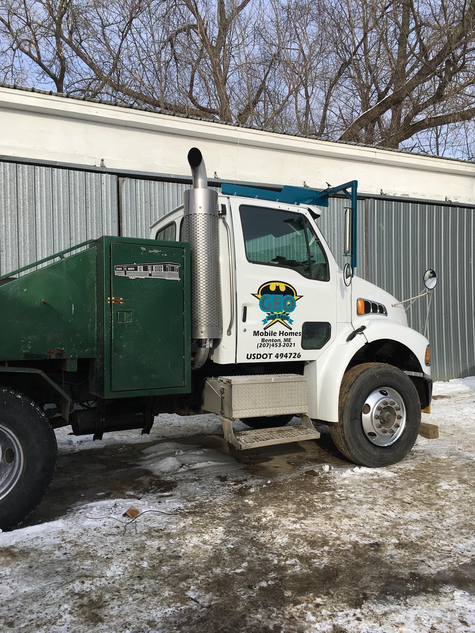 White and green dump truck parked in front of a metal building on a snowy day.