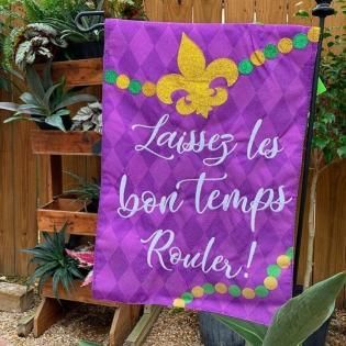 Purple sign with gold fleur-de-lis reading “Laissez les bon temps Rouler!” hanging on a wooden fence