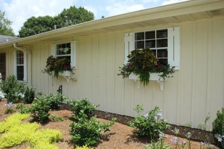 Single-story beige house exterior with white-trimmed windows, hanging flower baskets, and landscaped front garden.