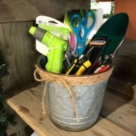 Metal bucket holding garden tools, including a green sprayer, hand trowel, and gloves, on a wooden floor.