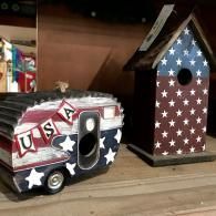 Two patriotic birdhouses on a shelf, one red-white-blue trailer and one red-white-blue birdhouse with stars.