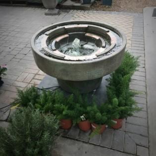 Small round stone water fountain with plants around it on a paved patio