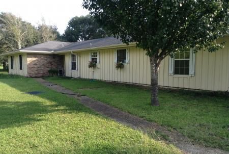 Single-story tan house with a brick entry, front lawn, and a tree-lined walkway