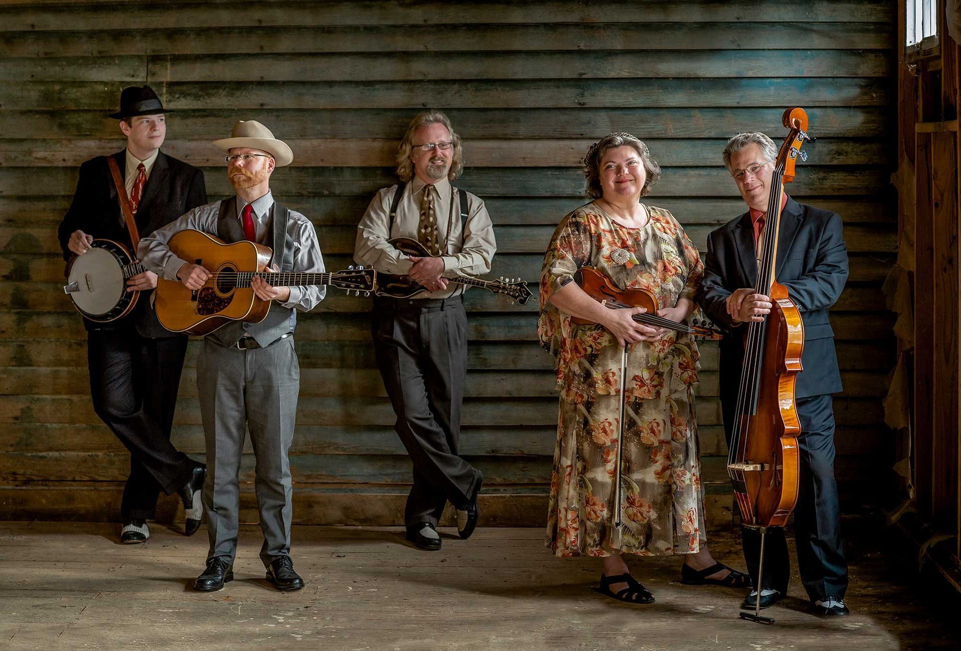 Five musicians pose against a weathered wooden wall. Instruments include banjo, guitar, mandolin, ukulele, and upright bass.