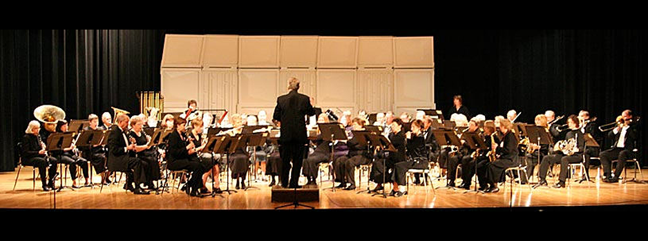 Park Rapids Area Community Band performing on a stage. The conductor faces the orchestra. Black background with tan backdrop.