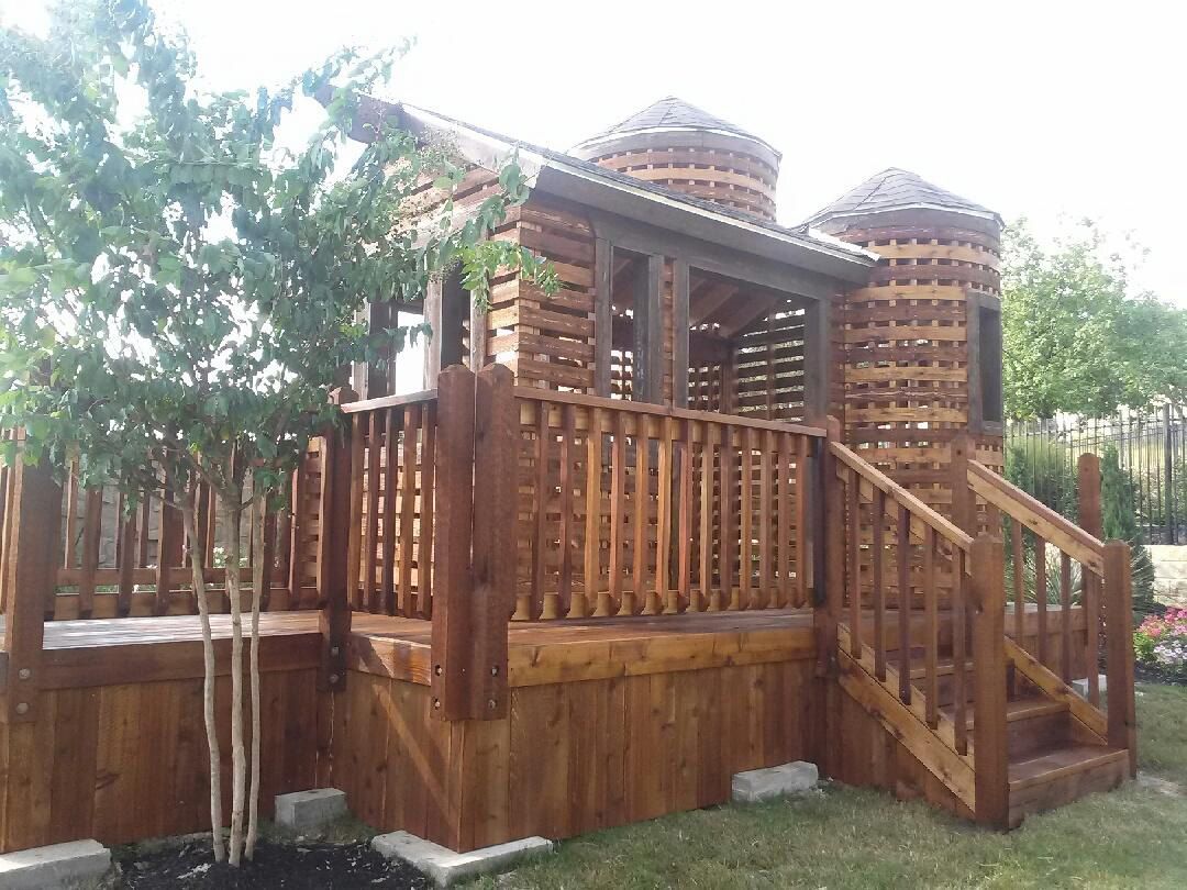 Wooden playhouse with a deck, stairs, and two silo-shaped towers, stained brown, on a grassy lawn.