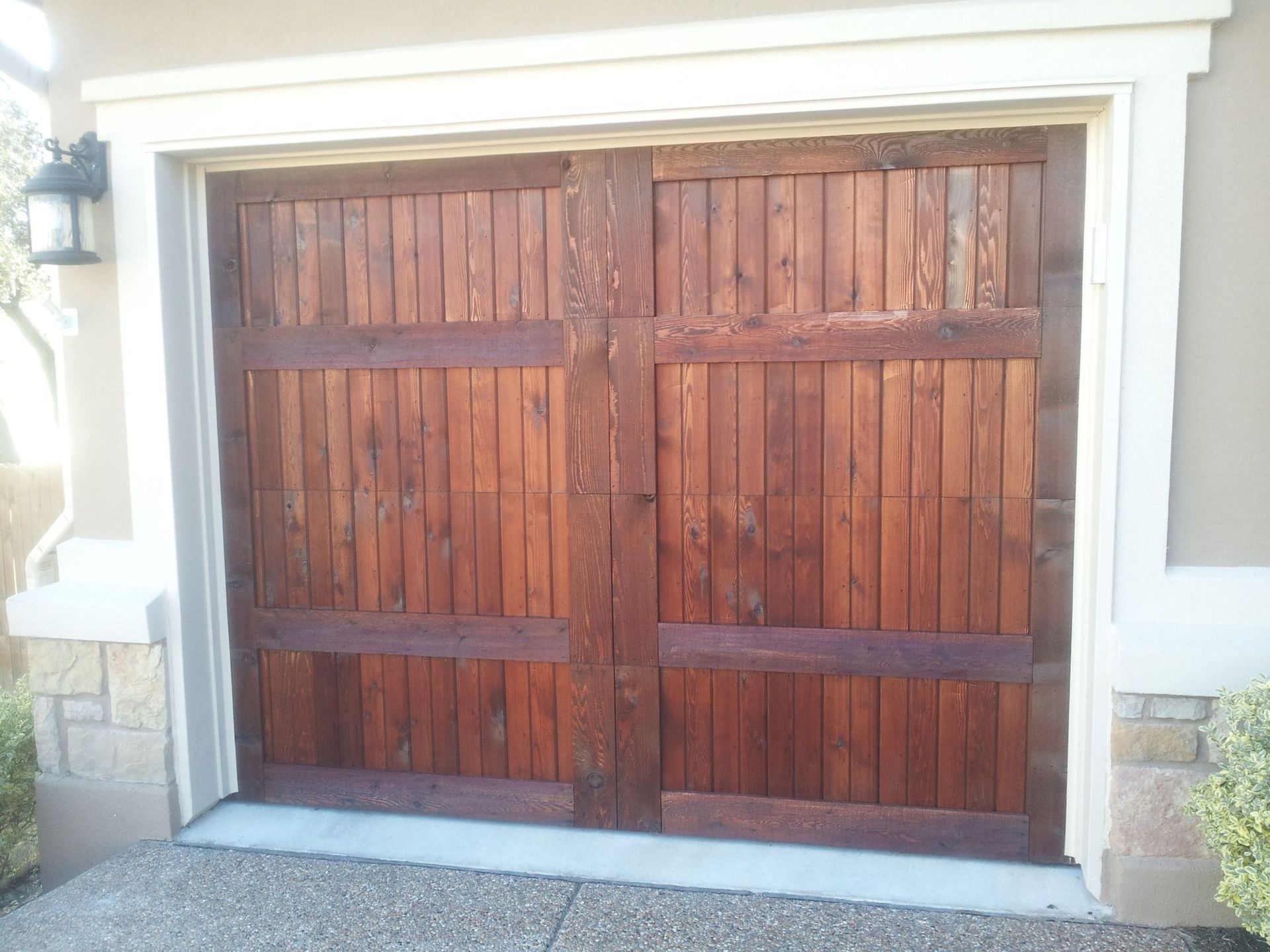 Wooden garage door with vertical planks and horizontal crossbars, framed in white trim, set in a stucco and stone facade.