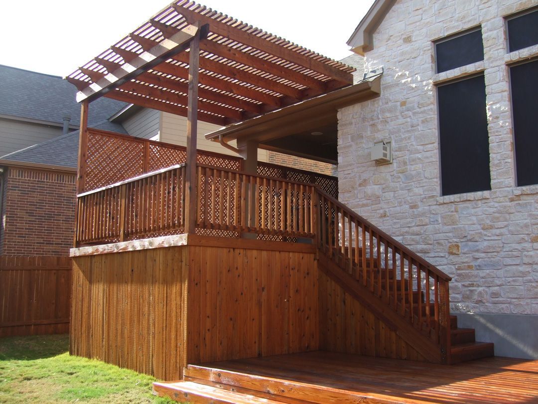 Wooden deck with pergola attached to a stone-clad house. Steps lead down to the yard.