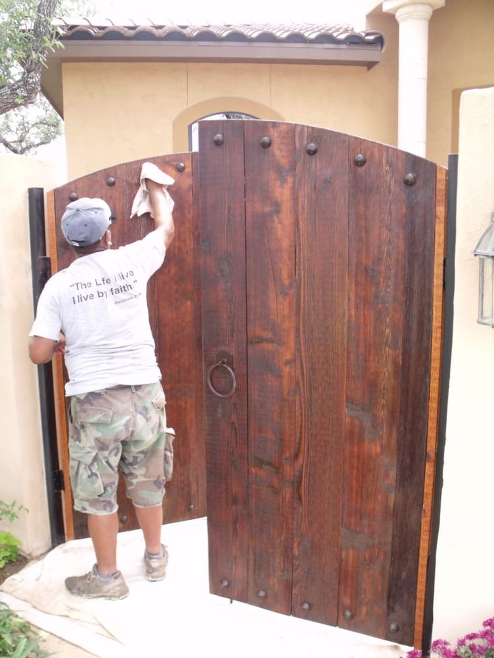 A person wiping down a dark stained wooden gate. The gate is brown with large metal studs, set in front of a beige wall.