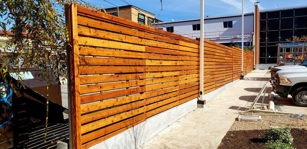 Horizontal wooden fence along a paved area with buildings in the background and a parked truck.