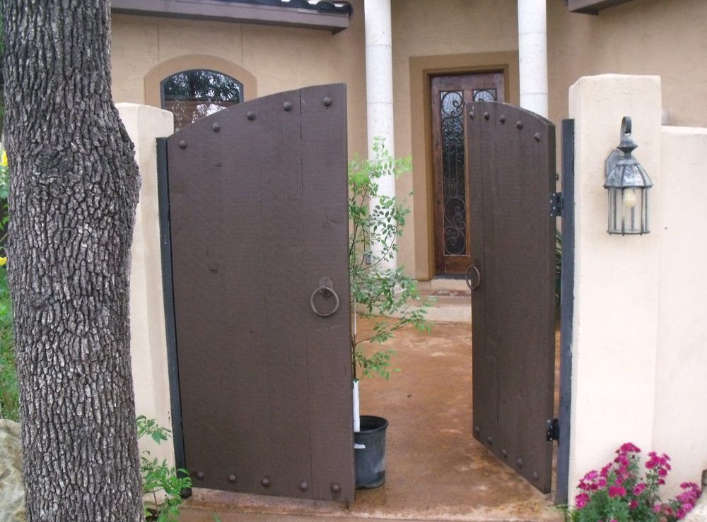 Open brown wooden gate in front of a tan-colored building with a door, light and flowers.
