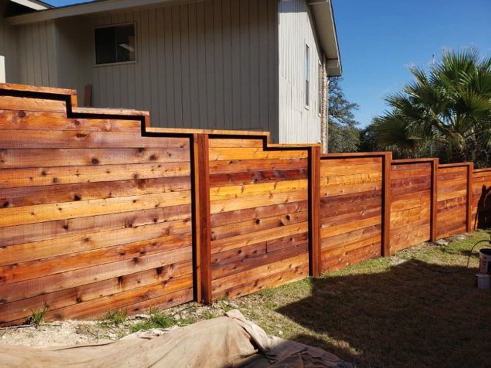 Wooden fence with varying heights, stained in shades of brown, next to a building on a sunny day.
