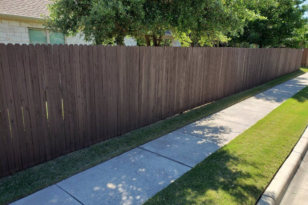 Brown wooden fence alongside a sidewalk and grassy strip, with trees in the background.
