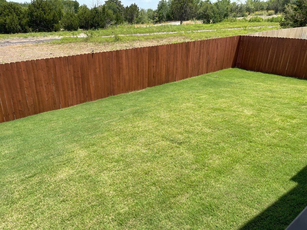 A rectangular grassy backyard enclosed by a brown wooden fence under a bright sky.