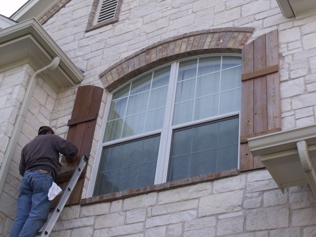 Man on ladder cleaning a window with shutters on a stone house.