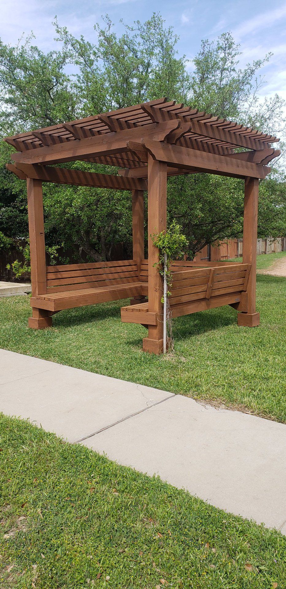 Wooden pergola with built-in benches in a grassy area next to a sidewalk.