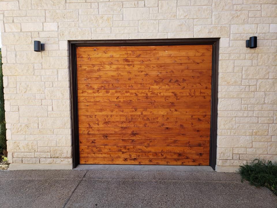 Wooden garage door with brown trim, tan stone wall, and two black sconces.