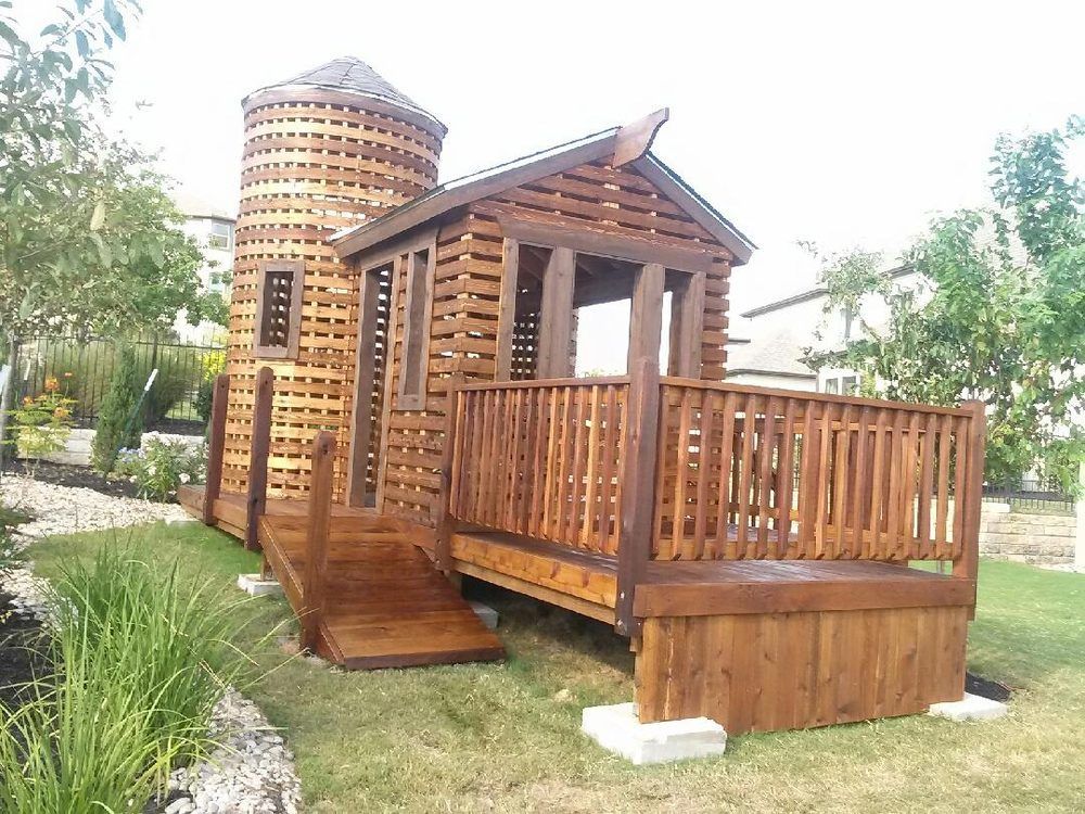 Wooden playhouse with a silo-like tower, deck, and ramp in a yard.
