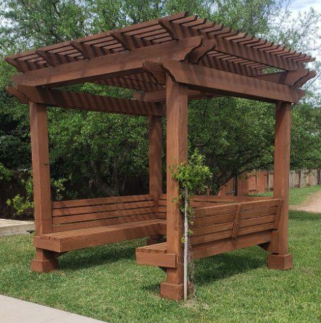 Brown wooden pergola with built-in benches, set in a grassy area with trees and a sidewalk.