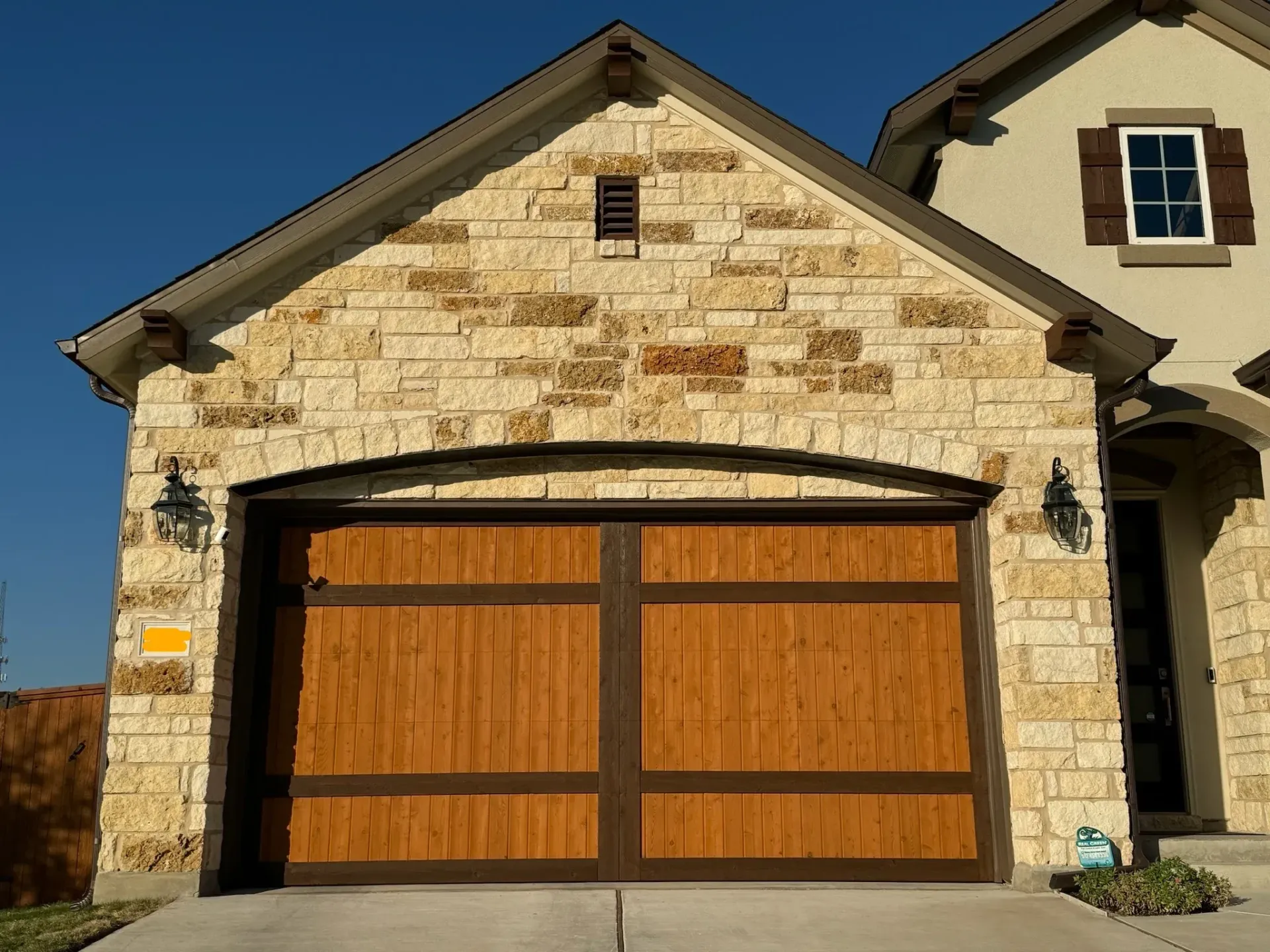 Tan stone garage with arched dark brown wood door, brown trim, and sconces.
